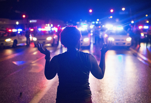 2015: A woman stands before police with her hands up in the air.