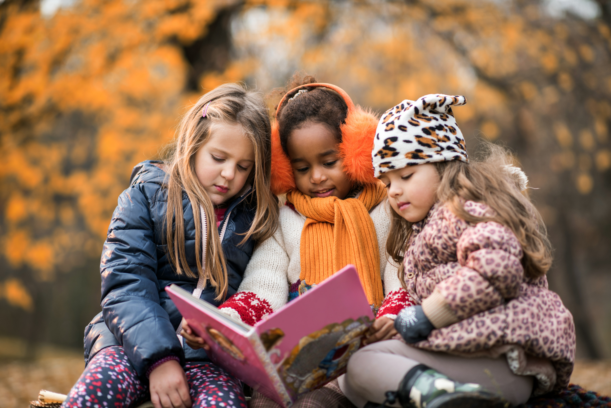 Three small girls reading a children's book in autumn day.