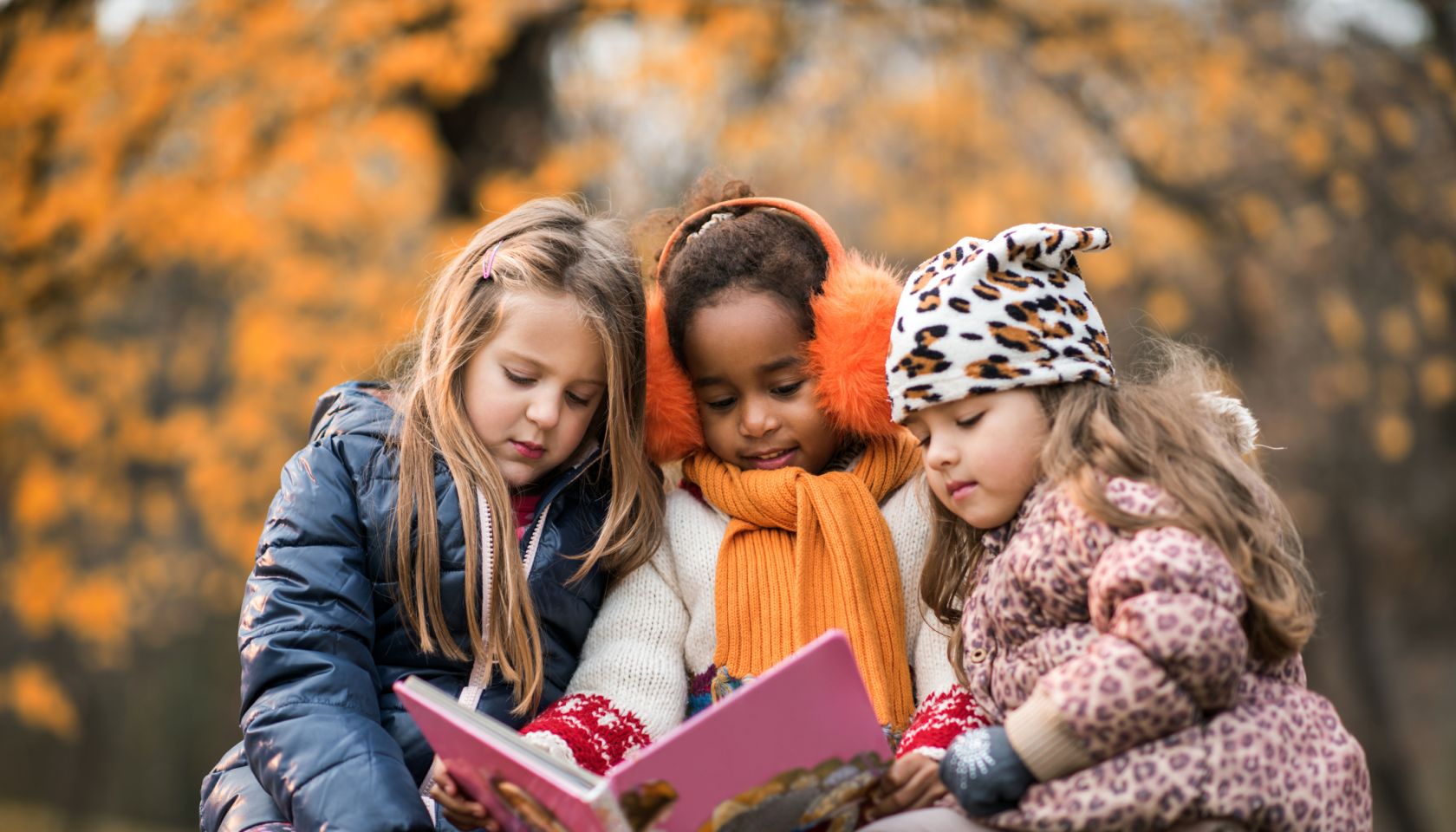 Three small girls reading a children's book in autumn day.