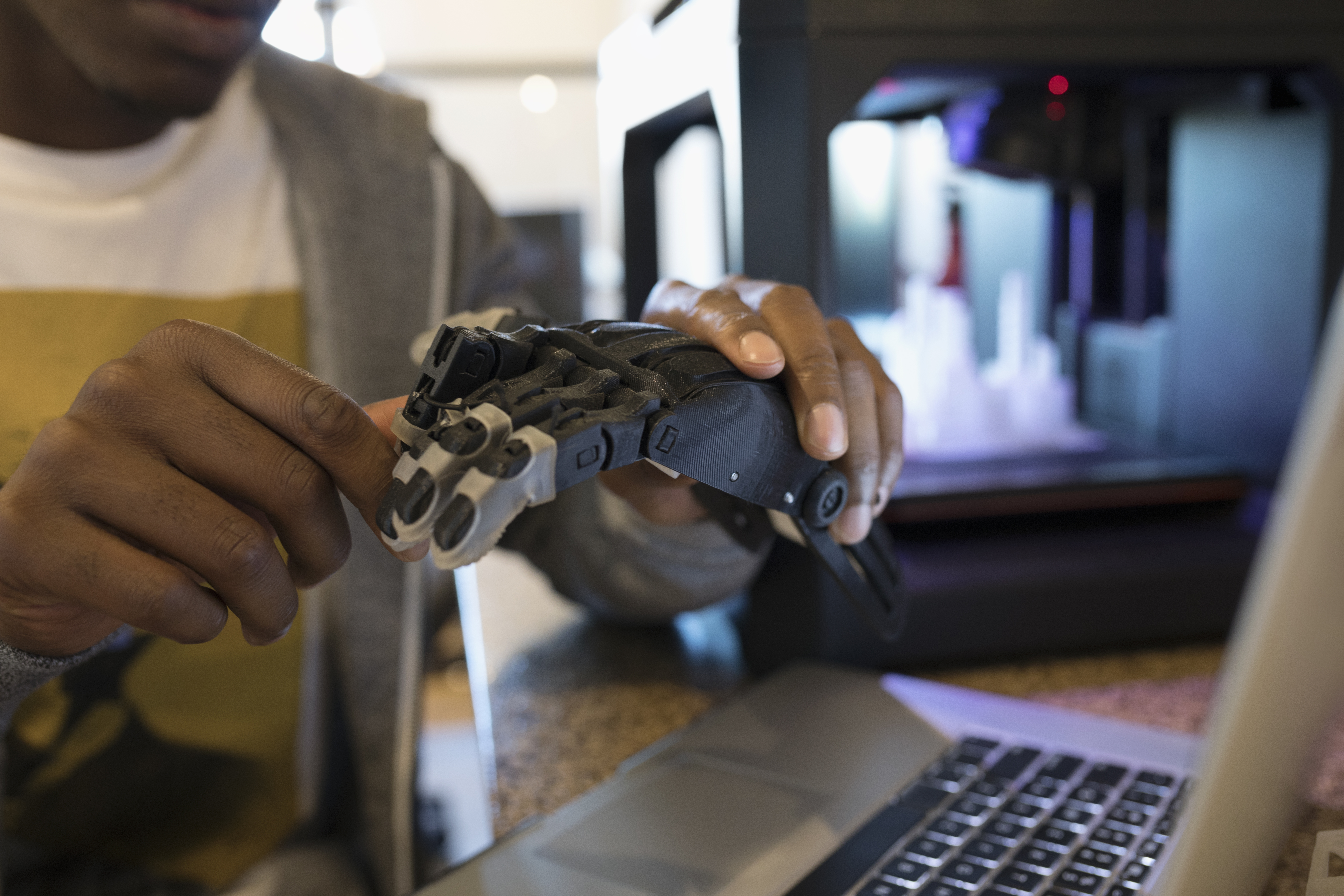 Close up male designer examining robotic hand prototype at laptop next to 3D printer