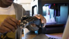 Close up male designer examining robotic hand prototype at laptop next to 3D printer