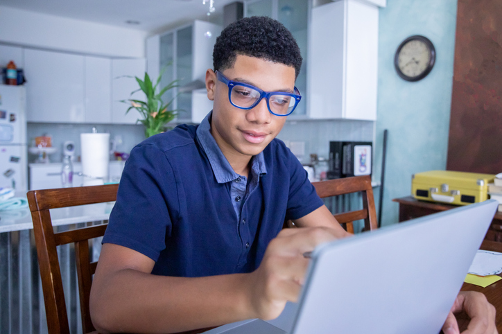 Smart teenage boy uses laptop computer to do homework in kitchen