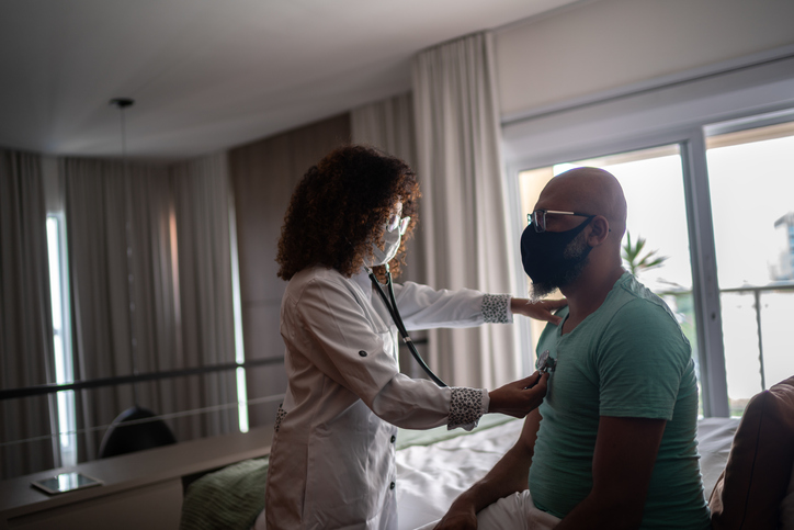 Doctor listening to patient's heartbeat during home visit - wearing face mask