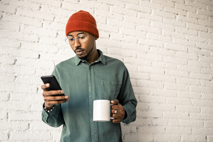 Man with coffee cup using smart phone against white brick wall