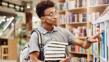 Serious Afro-American high school student in eyeglasses wearing satchel on one shoulder standing in modern library and finding books to prepare for exam