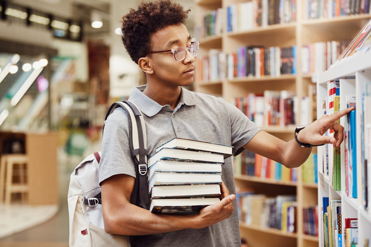 Serious Afro-American high school student in eyeglasses wearing satchel on one shoulder standing in modern library and finding books to prepare for exam
