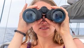 Young woman looking through binoculars, on sailboat, Dodecanese, Greece