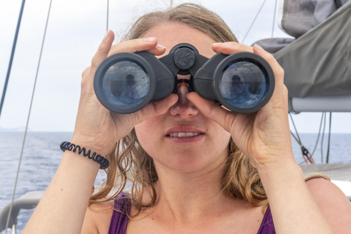 Young woman looking through binoculars, on sailboat, Dodecanese, Greece