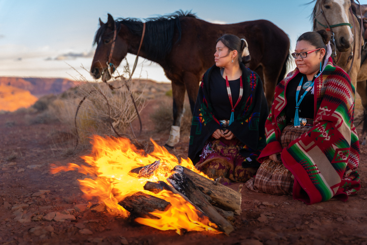 Two Native American Sisters, One Playing A Drum, Wrapped In Traditional Navajo Blankets Staying Warm By The Campfire Horses Behind Them, Iconic Monuments, Sunset