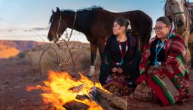 Two Native American Sisters, One Playing A Drum, Wrapped In Traditional Navajo Blankets Staying Warm By The Campfire Horses Behind Them, Iconic Monuments, Sunset