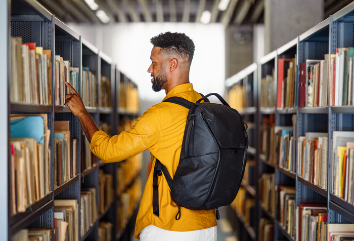 Rear view of young university student with book indoors in librabry.
