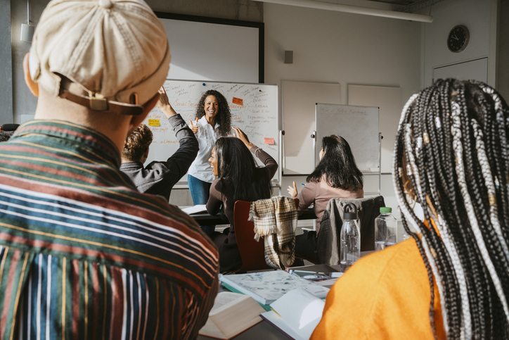 Happy professor looking at university students sitting with hands raised in classroom