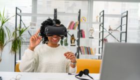 Virtual work.Young African woman in VR headset pointing in the air while sitting at the desk in creative office