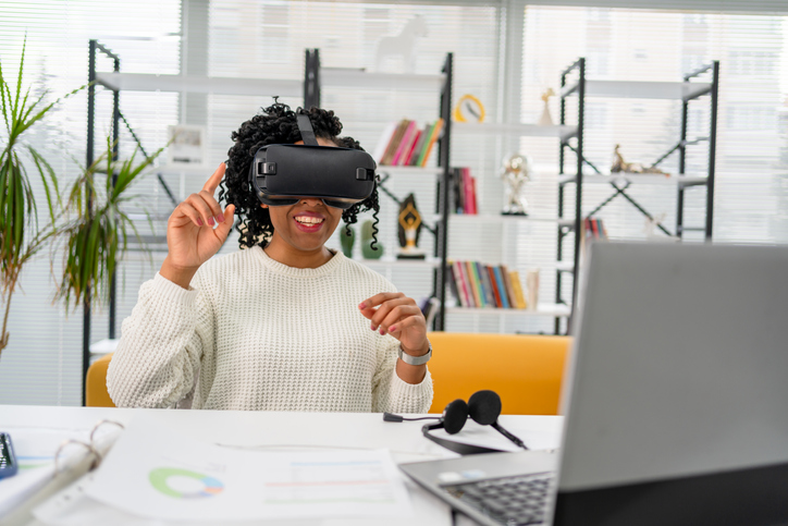 Virtual work.Young African woman in VR headset pointing in the air while sitting at the desk in creative office