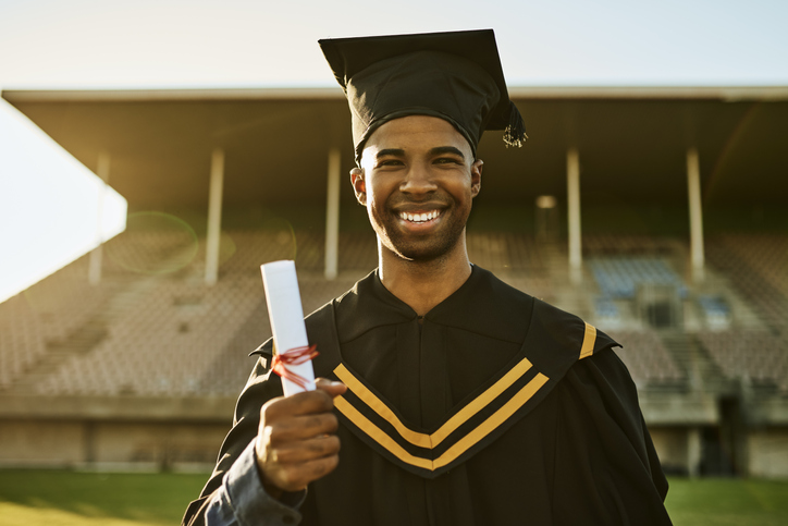 Portrait of smiling graduate in gown and hat standing alone on university campus at graduation ceremony. Excited, happy qualified postgrad graduating college for academic bachelor, masters and degree