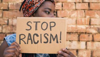 African american women standing in front of a brick wall, she is holding a sign that says Stop Racism