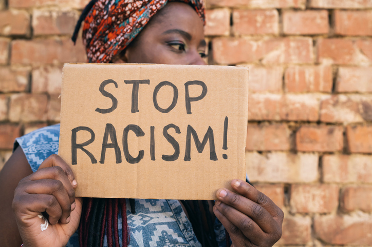 African american women standing in front of a brick wall, she is holding a sign that says Stop Racism