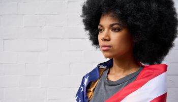 Serious African American girl wrapped in usa flag standing on white background.