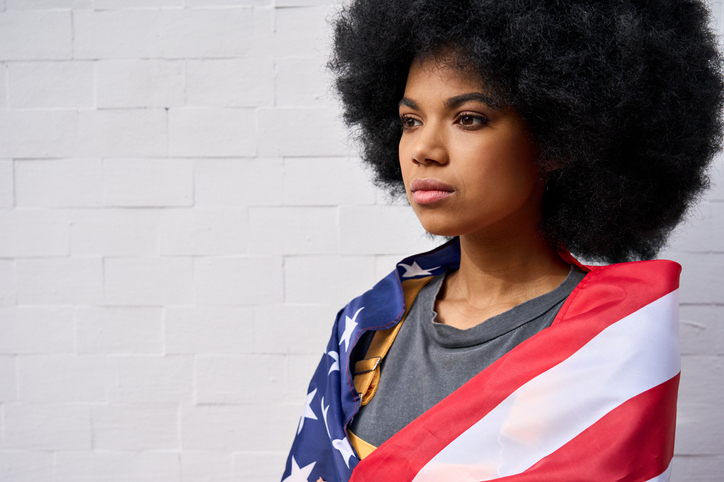 Serious African American girl wrapped in usa flag standing on white background.