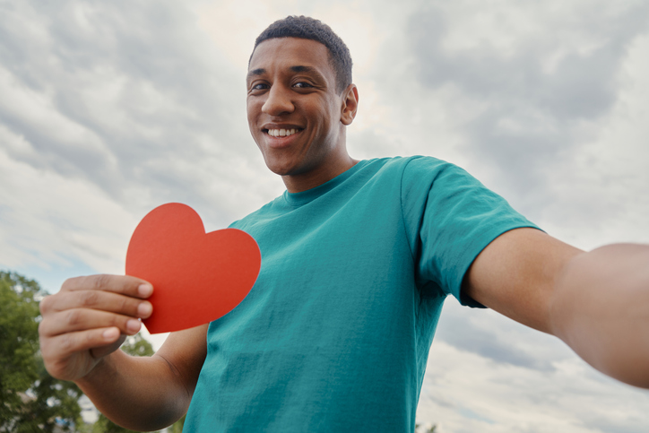 Happy mixed race man making selfie and holding paper heart shape with sky in the background