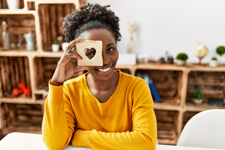 African american woman holding bread with heart hole shape over eye at home