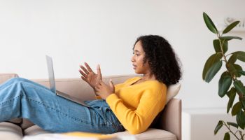 Positive young African American woman using laptop, relaxing on sofa at home, side view