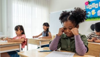 African American black girl student doing an exam at elementary school. Adorable young girl children sitting indoors on table, feeling upset and depressed while learning with teacher at kindergarten.