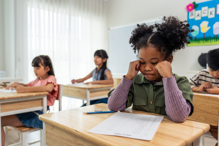 African American black girl student doing an exam at elementary school. Adorable young girl children sitting indoors on table, feeling upset and depressed while learning with teacher at kindergarten.