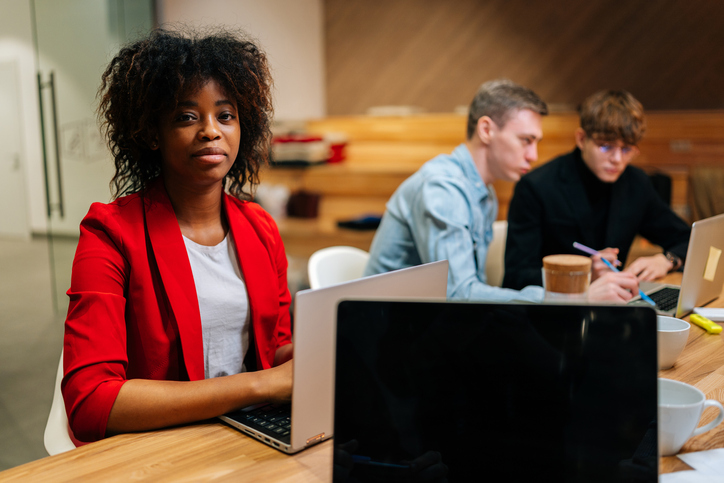 Portrait of happy African American businesswoman sitting at desk with laptop, looking at camera, during multiethnic professional businesspeople working together