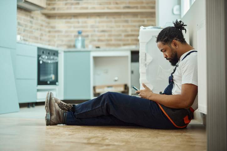 Black manual worker using cell phone on a break during home renovation process.