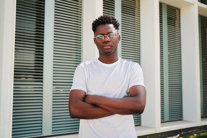 Portrait of young african american serious confident guy looking at camera with crossed arms standing outside. Handsome college student man posing with a calm pensive attitude at the university campus