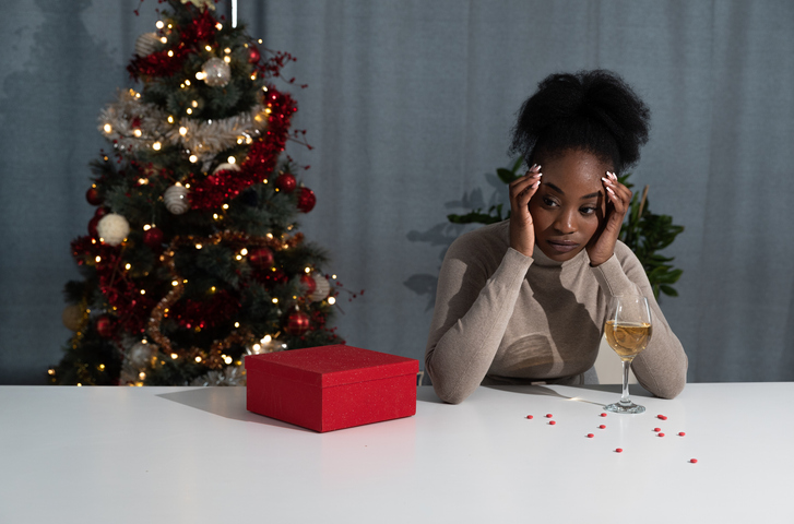 Young depressed and anxious African American woman sitting alone at home at Christmas Eve with alcohol drink and pain killers pills feeling desperate and hopeless. Holiday sadness concept