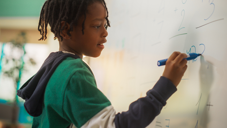 Portrait of a Handsome African Boy Finding a Solution to a Mathematical Task on a Whiteboard in Class in Elementary School. Young Pupil is Focused on Numbers, Writing Down a Correct Answer