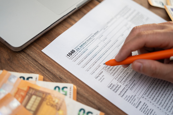 Close-up of a hand filling out a 1040 tax form with euros and a laptop on a wooden desk. Tax filing and financial preparation concept, ideal for accounting and international finance themes.