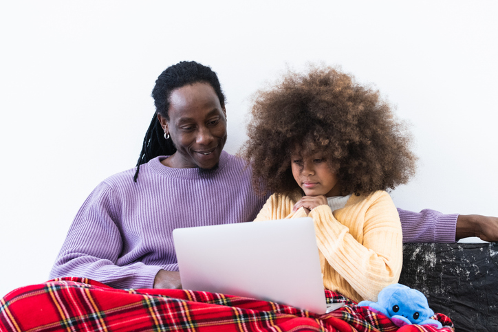 Black father browsing laptop with daughter under plaid