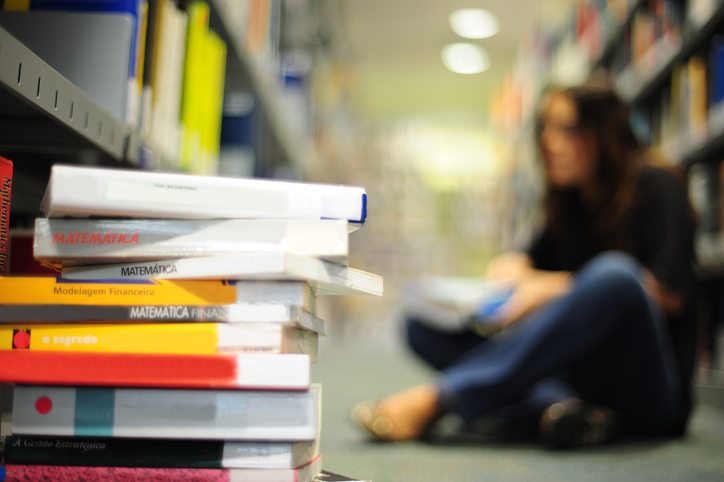Stack of Books in Focus with Blurred Person Sitting on Floor in Library Background