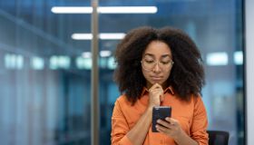 Serious thinking woman standing with phone in hands near window inside office. Businesswoman reading online news and browsing internet pages at workplace, using app