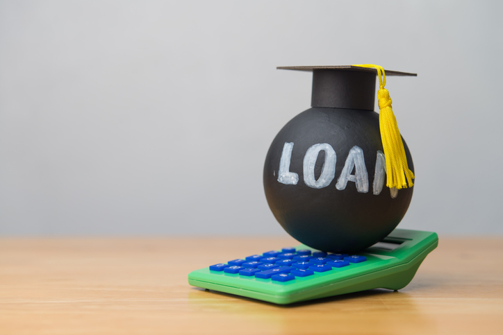 Heavy student loan wears mortarboard and green calculator on wooden table with white wall background.
