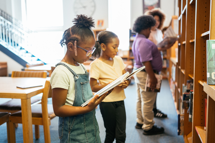 Diverse school girls with Latina teacher in library, picking up books from shelves and having a reading class