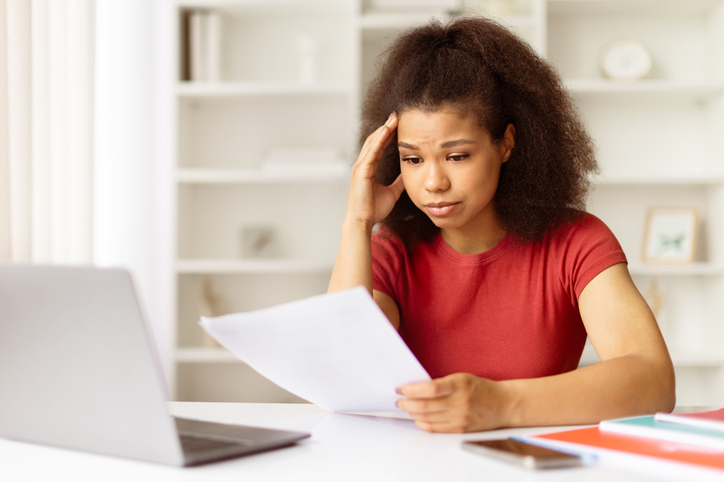 Young black woman looking stressed while reading documents at desk with laptop