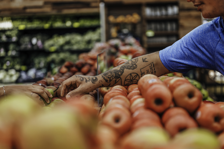 Tattooed Latino man reaching for an apple in the produce section of a grocery store
