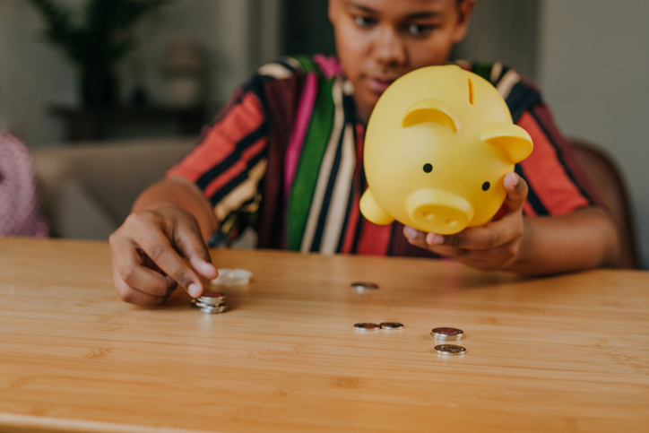 Teenage boy putting a coin into a piggy bank sitting at table at home
