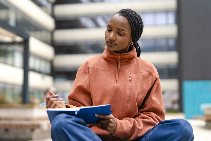 Young woman studying outdoors while writing in her notebook at a modern urban location
