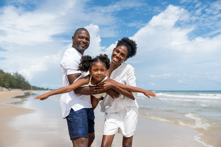 Happy black family travel enjoys holiday at beach with father and mother holding daughter in flying pose together under blue sky in summer