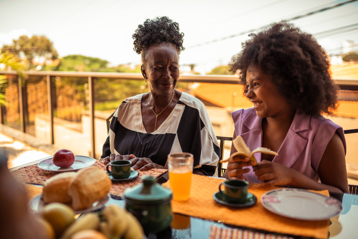 Happy African American grandmother and granddaughter enjoy a healthy breakfast together on a sunny outdoor balcony, sharing a joyful moment.