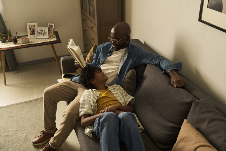 Father reading a book with son resting on sofa at home