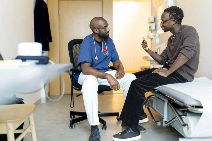 Full length of young man communicating problems through sign language to doctor while sitting on examination table in cl