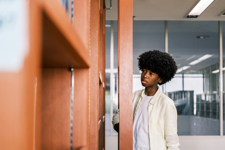A young student with curly hair searches for a book on a wooden shelf in a bright library, symbolizing learning, curiosity, and self-guided education in a diverse academic setting.