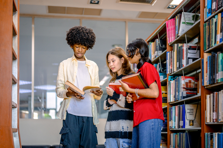 Three multiracial young friends discussing literature while standing in a modern school library aisle.
