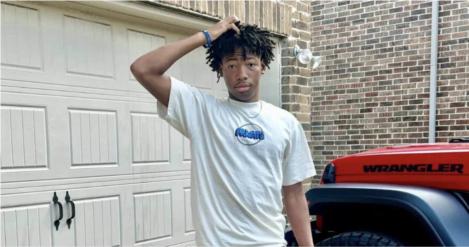 A young man with dreadlocks wearing a white t-shirt with a Pringles logo, standing in front of a red Jeep Wrangler.